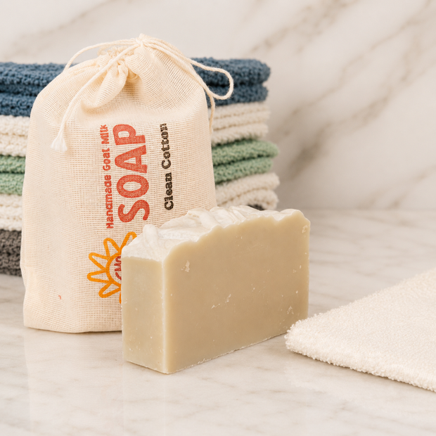Bar of goat milk soap with a drawstring bag labeled 'Soap' and folded towels in the background.