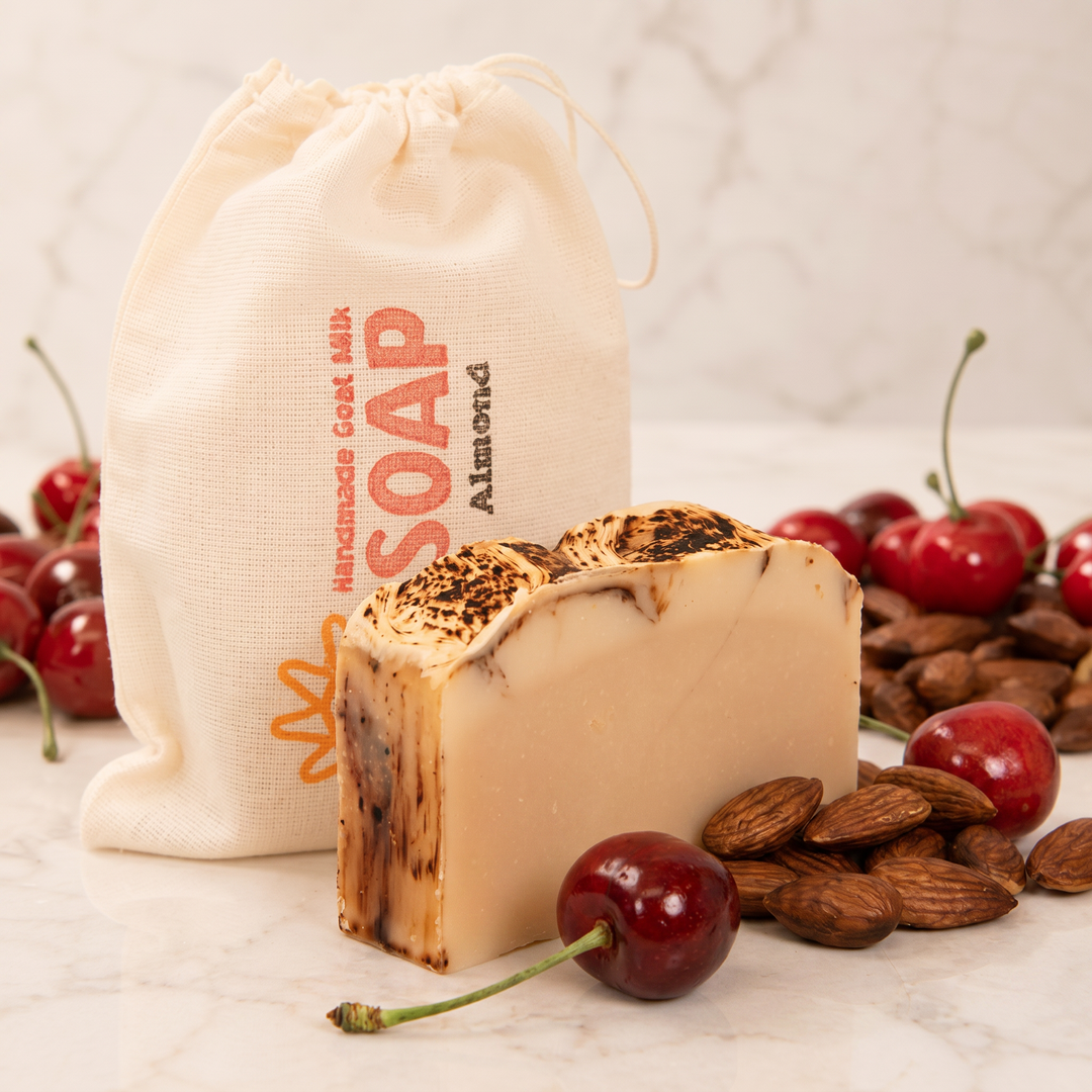 Bar of goat milk soap with almonds and cherries on a light surface, with a drawstring bag labeled 'Soap' in the background.