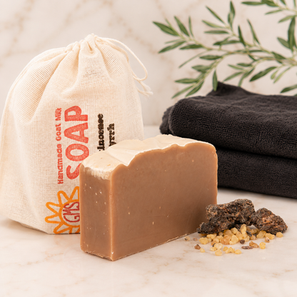Bar of goat milk soap with a drawstring bag labeled 'Handmade Soap' and black towels in the background.