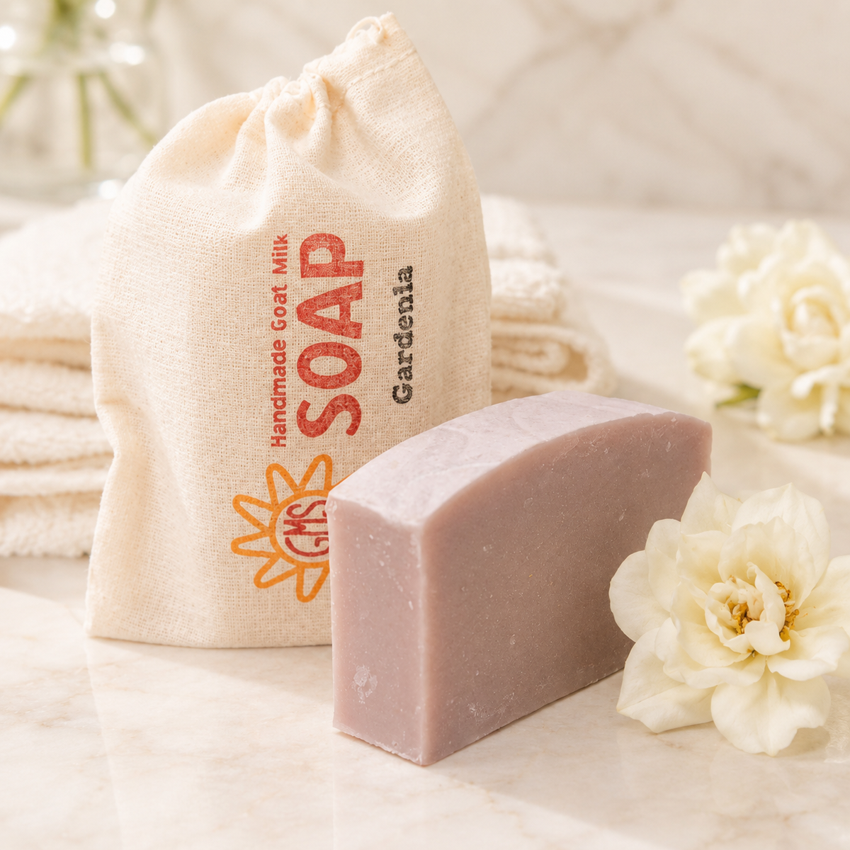 Bar of goat milk soap next to a drawstring bag labeled 'Handmade Goat Milk Soap' with 'Gardenia' on a light background.