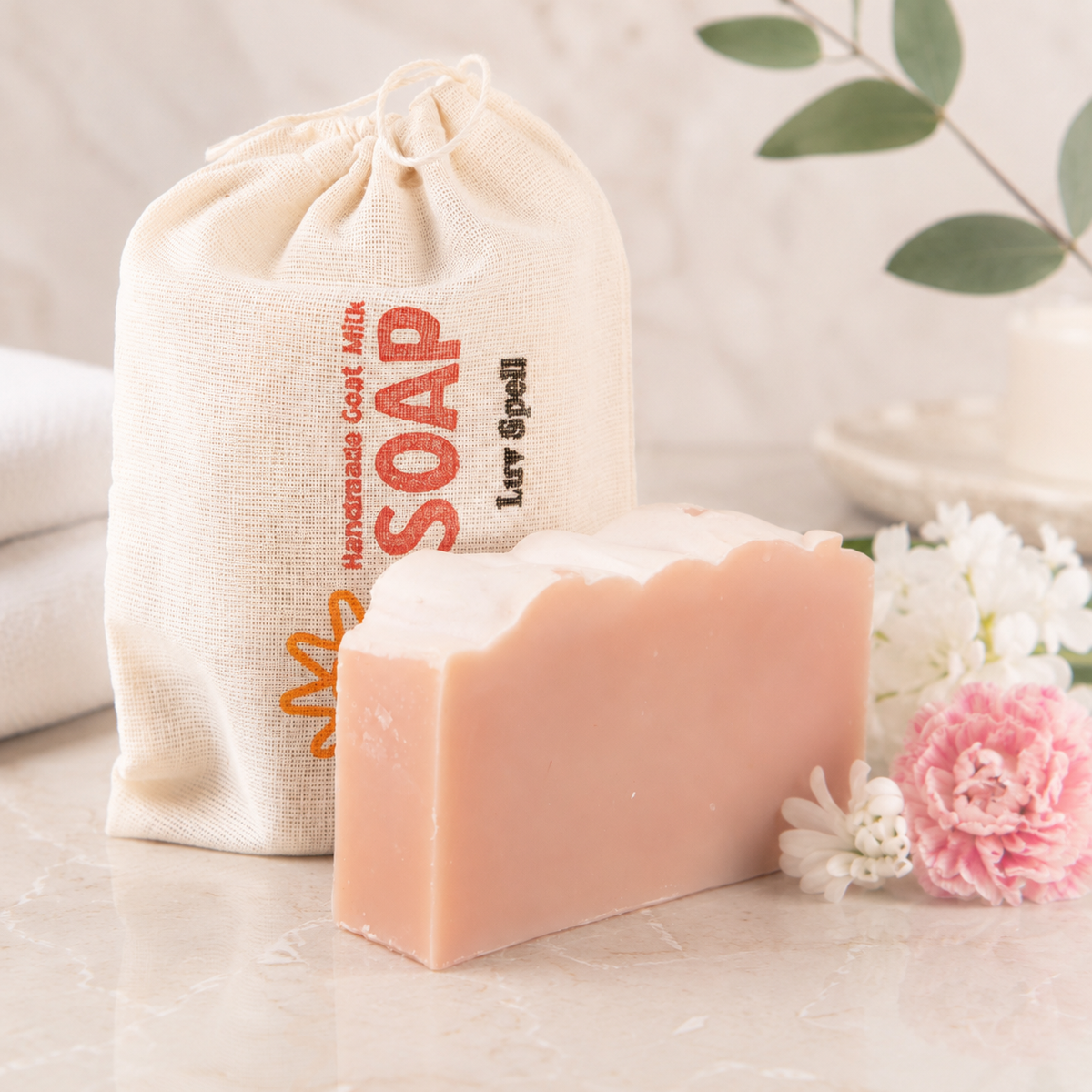 Bar of goat milk soap with a drawstring bag labeled 'SOAP' on a light surface with flowers.