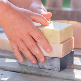 Someone holding a stack of three Goat Milk Soap bars on a wooden bench outdoors