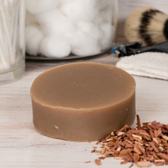 Cedarwood Shaving Goat Milk Soap resting on a wooden surface with a jar of cotton balls in the background and cedarwood beside it