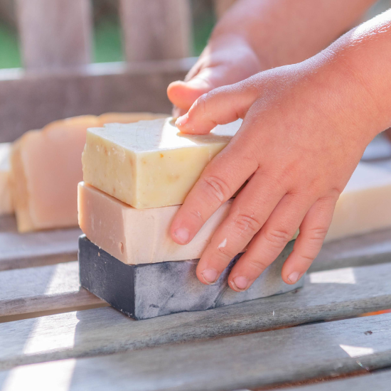 Kid's hand adjusting a stack of three Goat Milk Soap bars on a wooden surface outdoors