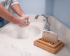 bar of soap sitting on wood soap dish with a person washing hands in the background