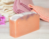 Rectangular goat milk soap violet bar on a textured surface with a pink fabric and flower in the background.